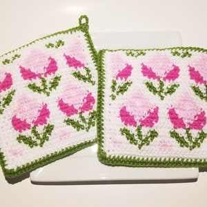 Könnte beinhalten: Zwei quadratische, weiße gehäkelte Topflappen mit Blumendesign. Die Blumen sind rosa und pink, mit grünen Stielen und Blättern. Die Ränder sind grün eingefasst. Jeder Topflappen hat eine kleine Schlaufe zum Aufhängen.