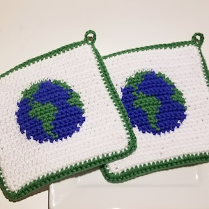 May include: Two square, white crocheted potholders with a blue and green Earth design in the center. The potholders have a green border and a loop for hanging. The potholders are handmade.