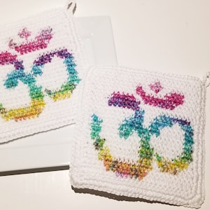May include: Two white, square crochet potholders with a colorful Om symbol. The Om symbol is in rainbow colors, and the potholders have a loop for hanging. The potholders are handmade and suitable for kitchen use.