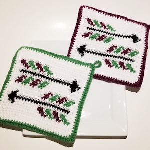 May include: Two crocheted pot holders with a white background and a design of black arrows with green and burgundy accents. The pot holders have a green and burgundy border.