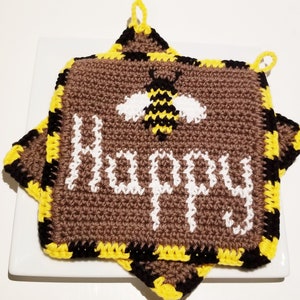 Puede incluir: Un agarrador de olla de color marrón tejido a crochet con un diseño de abeja negro y amarillo y la palabra "Happy" en blanco. El agarrador de olla tiene un lazo en cada esquina.