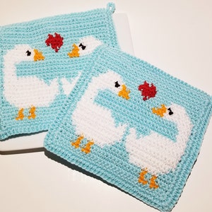 Puede incluir: Dos agarraderas de olla de ganchillo con fondo azul claro y gansos blancos con corazones rojos. Los gansos se miran y tienen picos y patas naranjas.