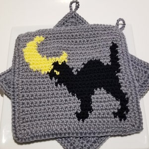 Op de afbeelding: Een paar grijze gehaakte ovenwanten met een zwart katten- en gele halve maanontwerp.