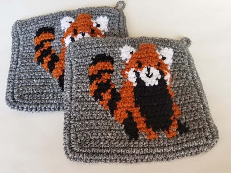 May include: Two gray crocheted pot holders with red panda designs. The red pandas are crocheted in orange, black, and white.
