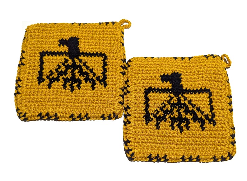 以下が含まれることがあります： マスタードイエローの正方形のかぎ針編み鍋つかみ2枚。中央に黒いワシのデザインがあります。縁は黒で縁取られ、それぞれに吊り下げ用の小さなループが付いています。手作りの鍋つかみです。