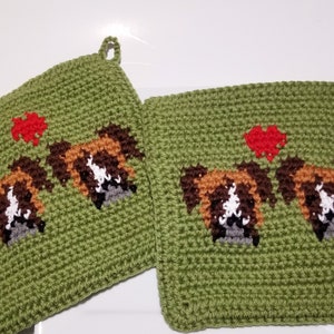 May include: Two green crocheted pot holders featuring a dog face design. The dog faces are brown, white, and grey, with a red heart above. Each pot holder has a hanging loop.
