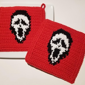 May include: Two red crocheted pot holders with a black and white ghost face design. Each square pot holder has a loop for hanging. The pot holders are displayed on a white plate.
