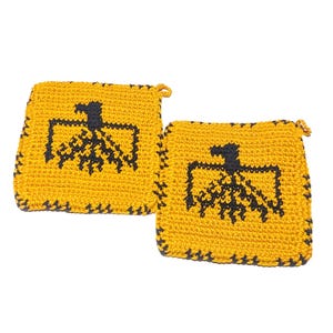以下が含まれることがあります： 2つの正方形の手作りのかぎ針編みの鍋つかみ。鍋つかみは黄色で、中央に黒の幾何学模様があります。エッジも黒です。各鍋つかみには、吊り下げ用の小さなループが付いています。