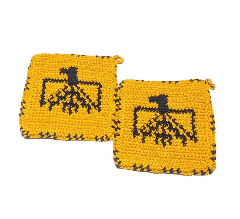 以下が含まれることがあります： 2つの正方形の手作りのかぎ針編みの鍋つかみ。鍋つかみは黄色で、中央に黒の幾何学模様があります。エッジも黒です。各鍋つかみには、吊り下げ用の小さなループが付いています。