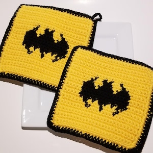 May include: Two yellow crocheted potholders with a black Batman logo. The potholders have a black border and a loop for hanging. They are square and sit on a white plate.