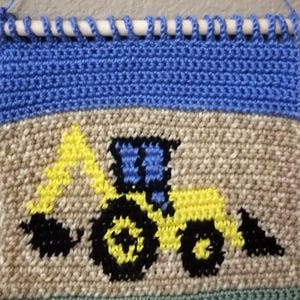 Könnte beinhalten: Handgehäkeltes Wandbehang mit einem gelb-blauen Traktormotiv. Der Traktor befindet sich auf einem beigefarbenen Hintergrund mit blauen und grünen Rändern. Er hängt an einem weißen Holzstab, befestigt mit blauem Garn. Ca. 30 cm breit.