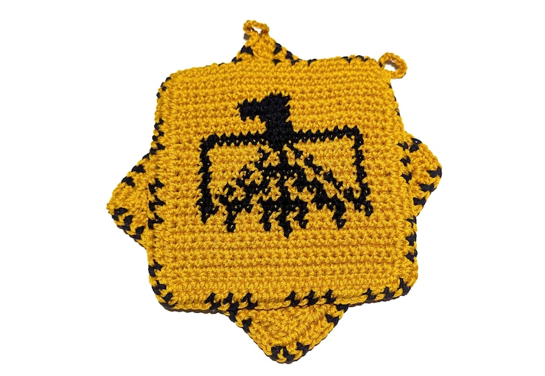 以下が含まれることがあります： 手作りの星型の編み物鍋つかみ。鍋つかみは黄色で、中央に黒いワシのデザインがあります。縁は黒で縁取られています。吊り下げ用のループが付いています。キッチンアクセサリーです。