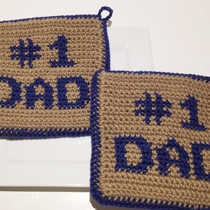 Op de afbeelding: Twee gebreide ovenwanten met een bruine achtergrond en blauwe stiksels. De tekst "#1 Dad" is in blauw gestikt op elke ovenwant.