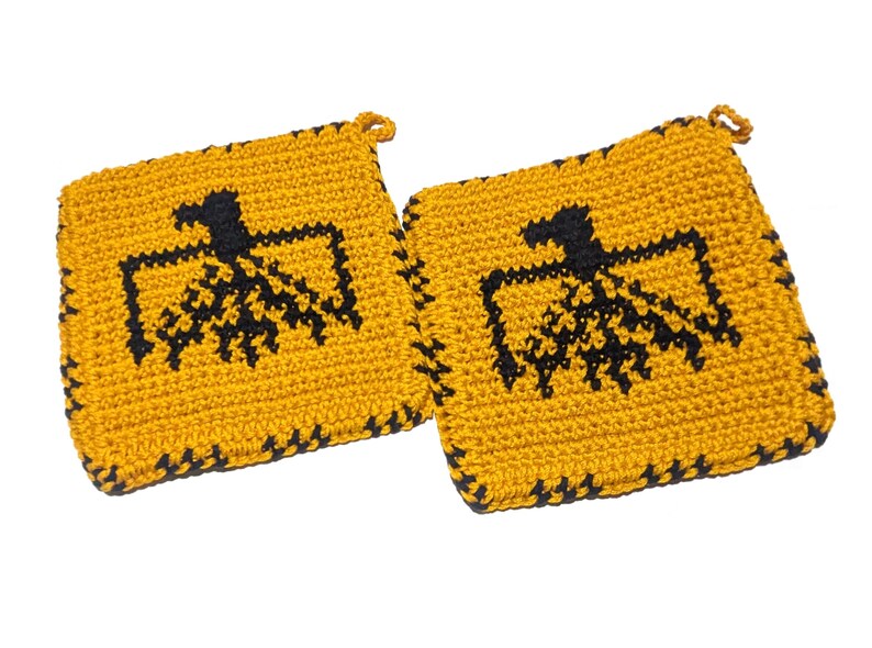 以下が含まれることがあります： 2つの正方形のかぎ針編み鍋つかみ。鍋つかみは黄色で、中央に黒い鳥のデザインがあります。縁は黒で縁取られています。各鍋つかみには、吊り下げ用の小さなループが付いています。
