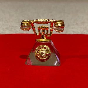 May include: A miniature gold-colored telephone with a clear, faceted base. The phone features a classic design with a handset, dial, and ornate detailing. It sits on a bright red surface, creating a striking contrast.