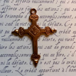 May include: A detailed, antique-style bronze cross with ornate detailing. The cross is centered against a backdrop of handwritten script in faded ink. The cross has a loop at the top for hanging.