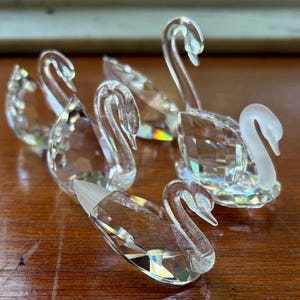 May include: A collection of clear crystal swan figurines. Each swan has a curved neck and detailed wings, reflecting light. The swans are arranged on a wooden surface, showcasing their intricate design and sparkling facets.