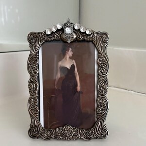 May include: A rectangular picture frame with an ornate, dark silver-toned metal border featuring embossed scrollwork and floral motifs. The top center is adorned with a cluster of clear, faceted crystals. It holds a portrait of a person in a black dress.