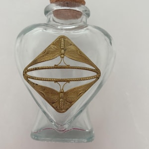 May include: A clear glass bottle shaped like a heart with a cork stopper. The front of the bottle features a gold-toned metal overlay in a diamond shape, decorated with two symmetrical butterfly motifs and a central horizontal oval frame.
