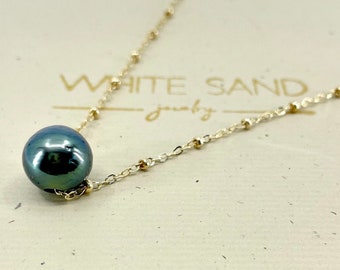 May include: A dark green pearl necklace with a gold chain and a small arrow charm. The necklace is in a white box with the words "White Sand Jewelry" printed on the top.