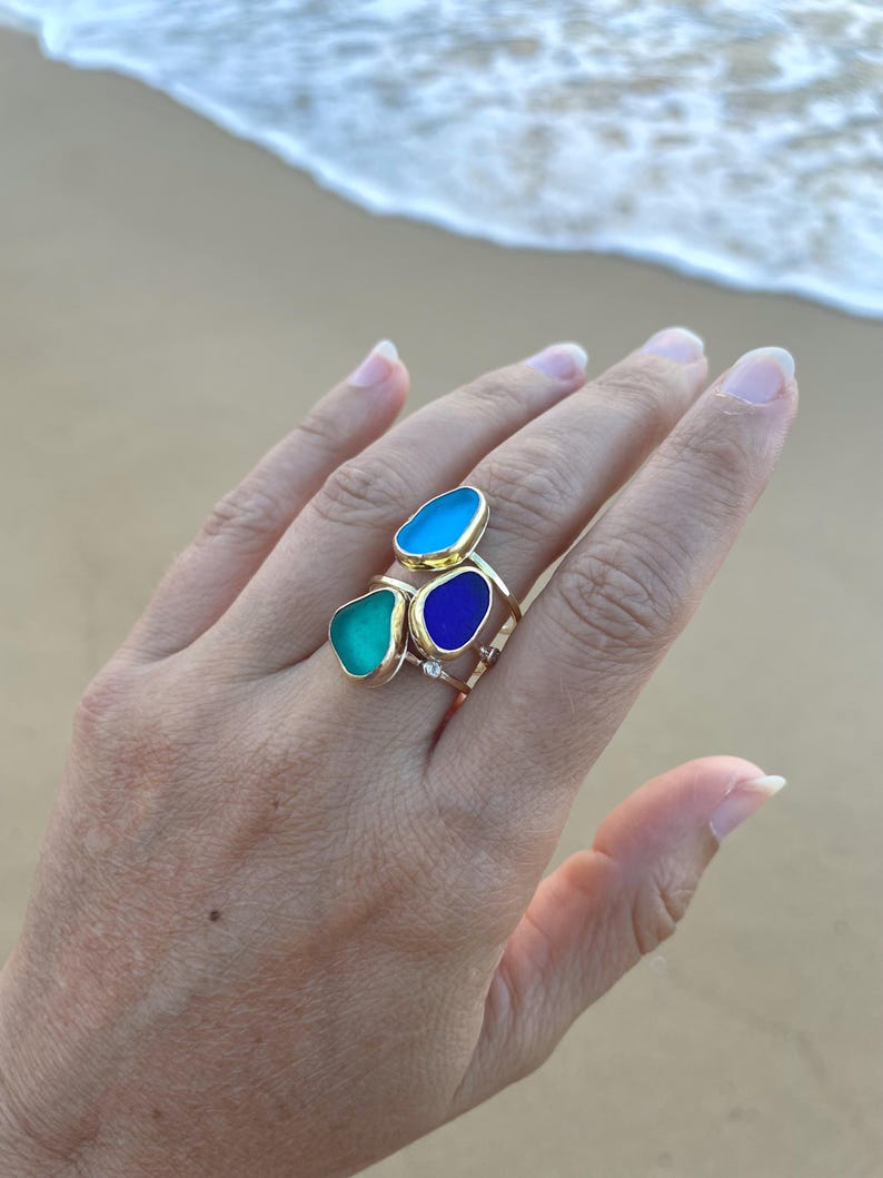 May include: Three gold rings with different coloured glass stones. The stones are turquoise, blue, and a darker blue. The rings are stacked on a finger.