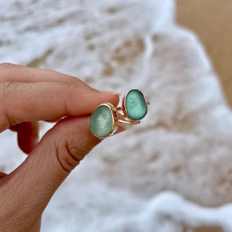 May include: Two rings featuring a light green sea glass stone set in a gold band. The rings are on a finger.
