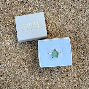 May include: A green sea glass ring in a white box with the text "WHITE SAND" on the lid.