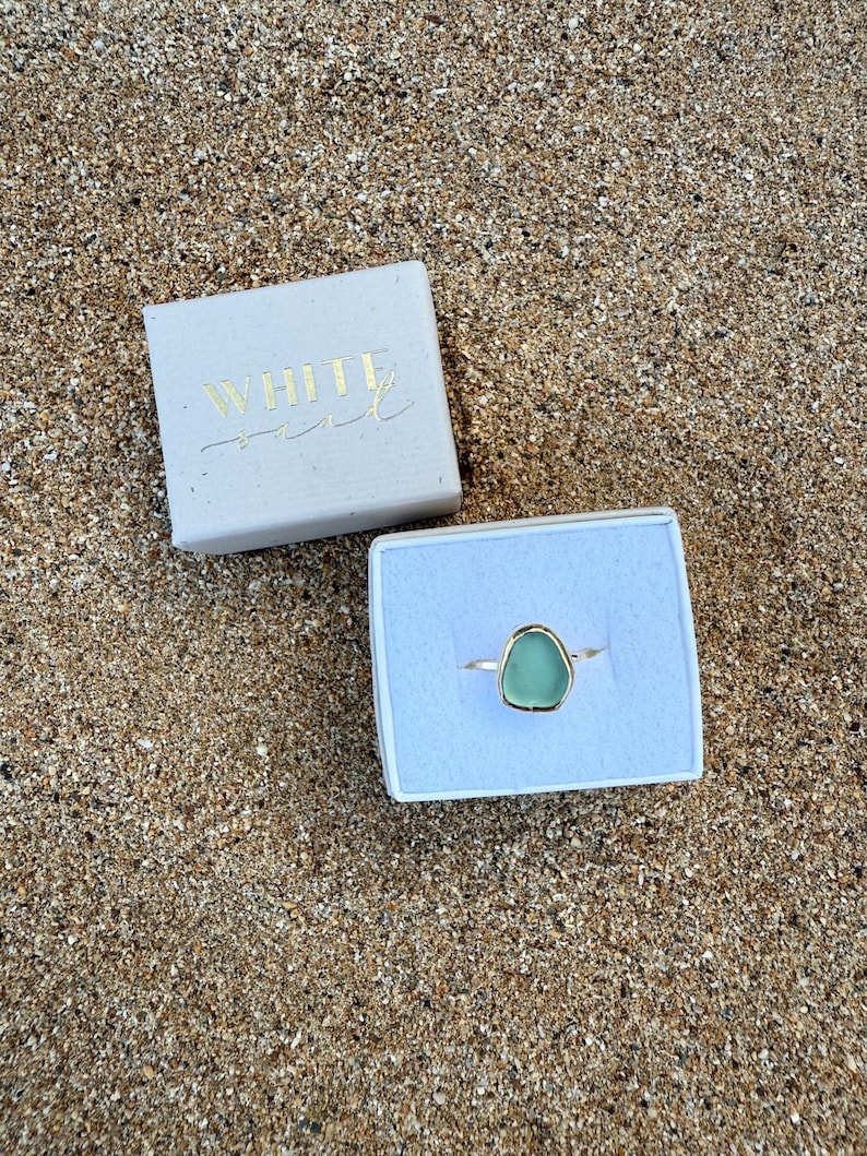 May include: A green sea glass ring in a white box with the text "WHITE SAND" on the lid.