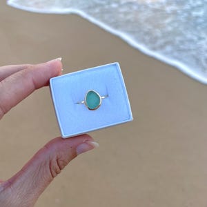 May include: A teardrop-shaped ring with a light blue gemstone set in a gold band. The ring is in a white box.