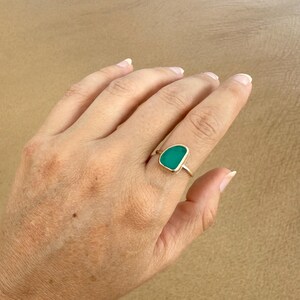 May include: A gold ring with a green sea glass stone. The ring is on a finger.