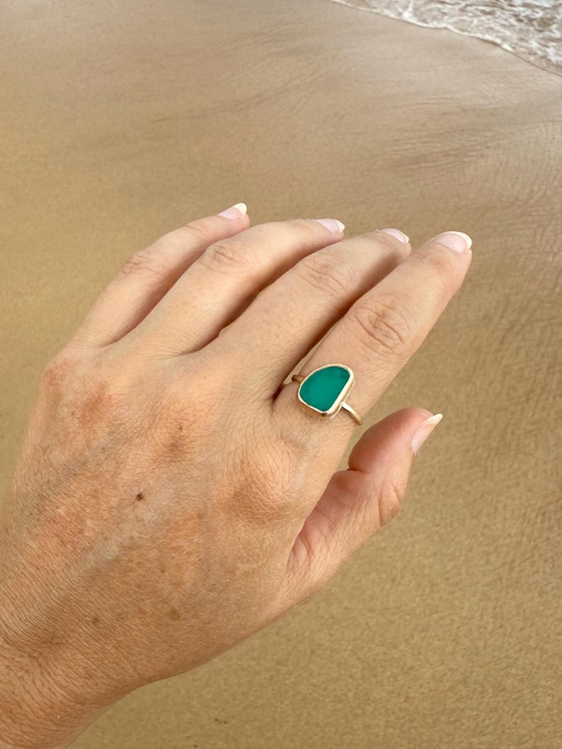 May include: A gold ring with a green sea glass stone. The ring is on a finger.