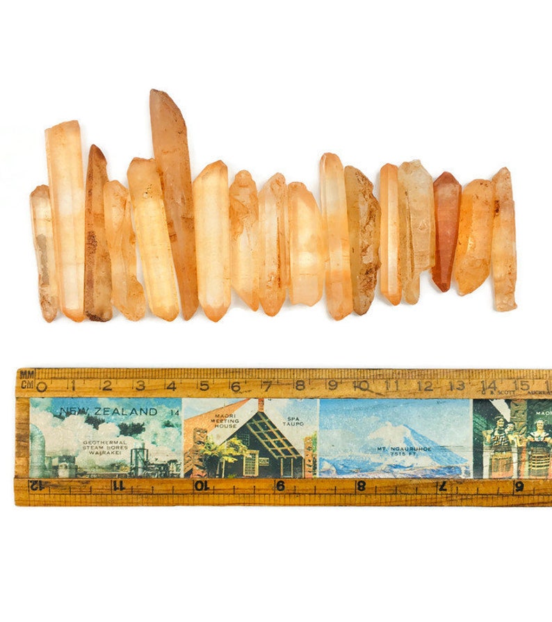 May include: A collection of 18 orange quartz crystal points, ranging in size from 1 to 2 inches, arranged in a row on a white surface. A ruler is placed below the crystals for scale.
