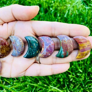 May include: A set of seven crescent moon shaped polished stones in various shades of red, brown, green, and white. The stones are arranged in a row, with the tips of the crescents facing the same direction.
