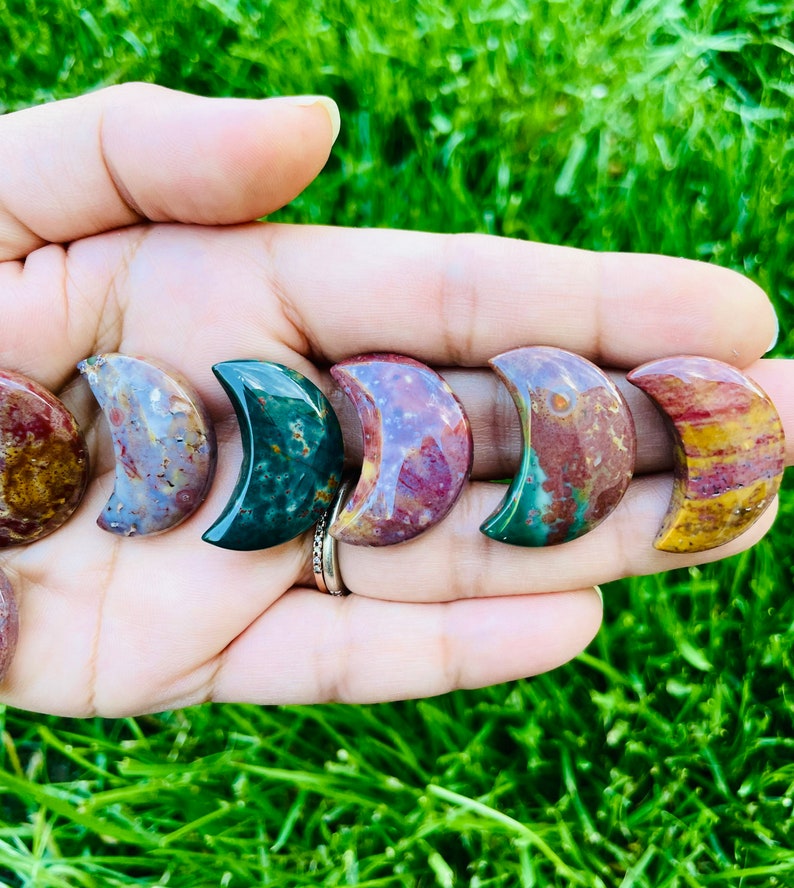 May include: A set of seven crescent moon shaped polished stones in various shades of red, brown, green, and white. The stones are arranged in a row, with the tips of the crescents facing the same direction.