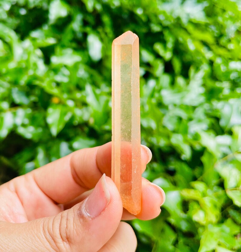 May include: A clear, orange-toned crystal point with a pointed tip. The crystal is translucent and has a slight pink hue.