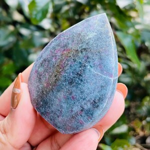 Puede incluir: Una piedra en forma de lágrima con un color base azul-gris y motas rojas y moradas. La piedra se sostiene en una mano, con un fondo verde borroso de follaje.