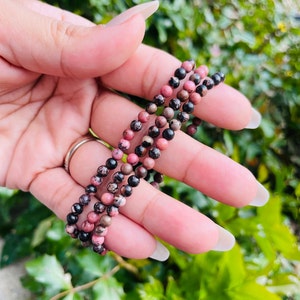 May include: A multi-strand bracelet made of pink and black gemstone beads. The beads are round and have a speckled pattern.