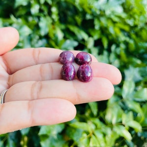 May include: Four cabochon ruby gemstones in a hand. The gemstones are a deep red color with a slight purple hue. They are smooth and polished.