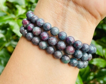May include: Three beaded bracelets with a mix of gray, teal, and red-purple gemstones. The round beads are strung together, creating a layered effect. The bracelets are worn on a wrist, with a blurred green background.