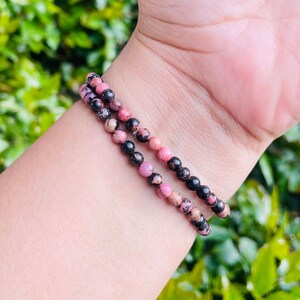 May include: A pink and black beaded bracelet with a stretchy band. The bracelet is made of small, round beads that are a mix of pink and black.