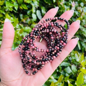 May include: A strand of pink and black gemstone beads. The beads are small and round, and they are strung together on a clear thread. The beads are arranged in a loose, flowing pattern.