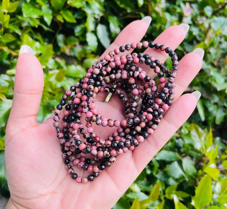 May include: A strand of pink and black gemstone beads. The beads are small and round, and they are strung together on a clear thread. The beads are arranged in a loose, flowing pattern.