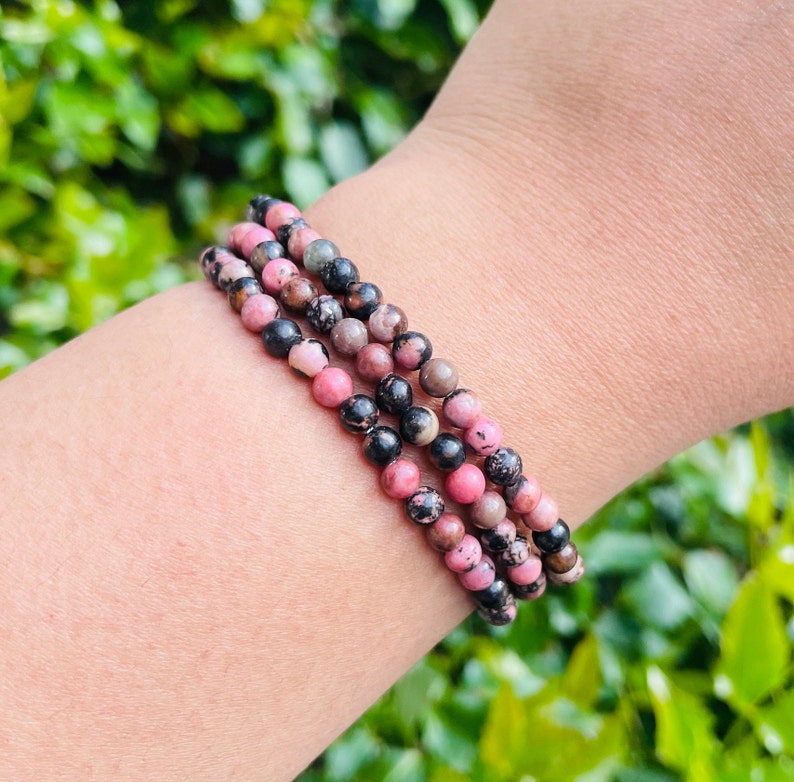 May include: A set of three bracelets made with pink and black patterned beads. The bracelets are stacked on a person's wrist.