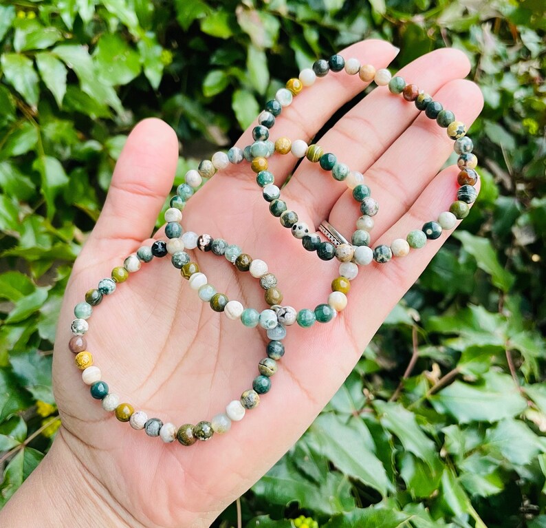 May include: Three green, brown, and white gemstone bracelets stacked on a hand. The bracelets are made of small, round beads.