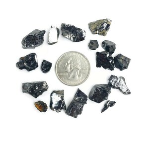 May include: A collection of black, metallic, and iridescent mineral specimens, some with a rough texture, scattered on a white surface. A US quarter is included for scale.