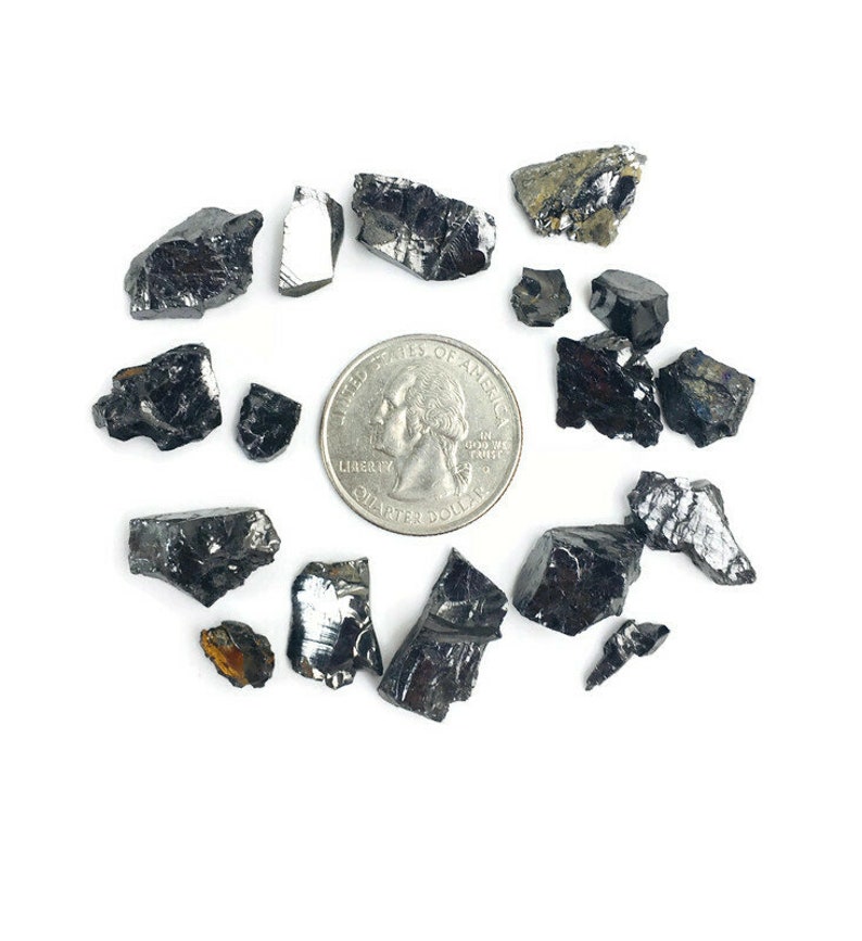 May include: A collection of black, metallic, and iridescent mineral specimens, some with a rough texture, scattered on a white surface. A US quarter is included for scale.