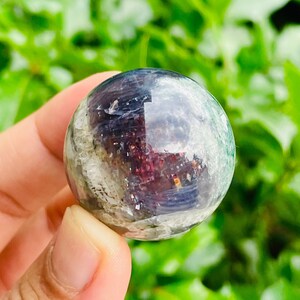 May include: A clear, spherical crystal ball with internal inclusions of purple, green, and white. The polished surface reflects light, creating a shimmering effect. The crystal is held in a hand against a blurred green background.