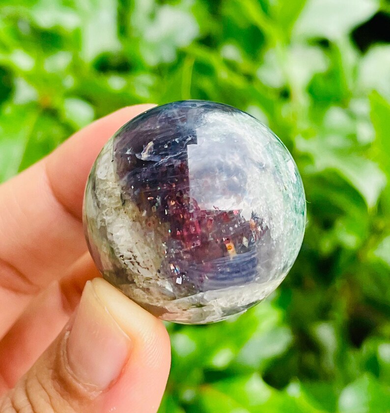 May include: A clear, spherical crystal ball with internal inclusions of purple, green, and white. The polished surface reflects light, creating a shimmering effect. The crystal is held in a hand against a blurred green background.