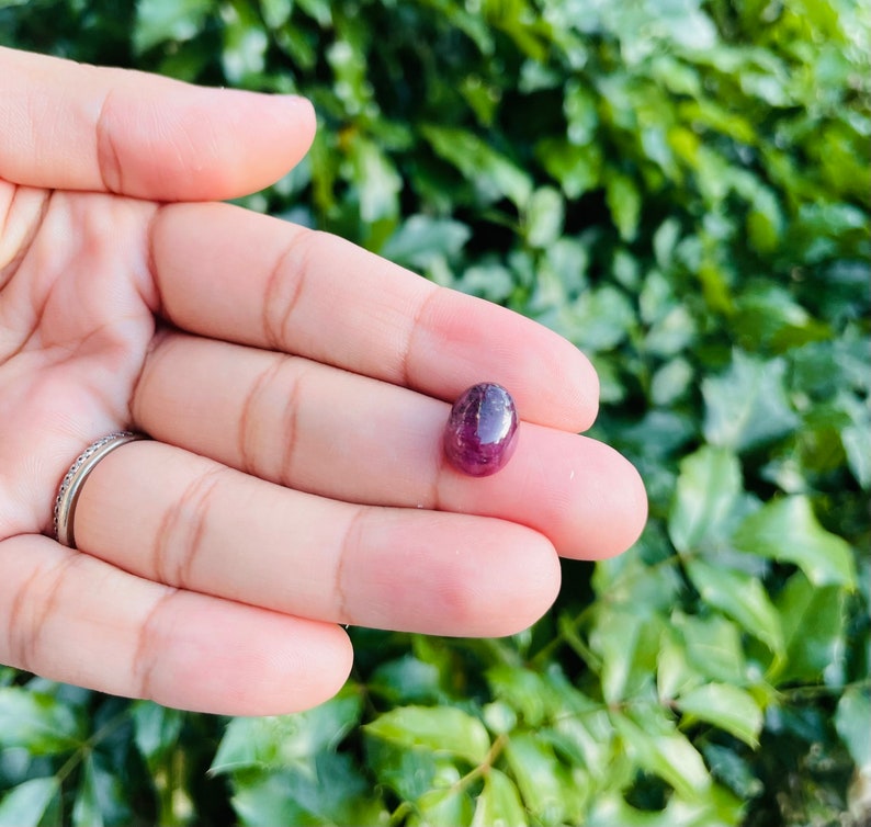 May include: A close-up of a deep red oval-shaped gemstone held in a hand. The gemstone has a slightly rough texture and is a rich, dark red color.