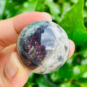 May include: A clear, spherical crystal ball with internal inclusions of dark red, green, and iridescent glitter. The sphere is held in a hand against a backdrop of green foliage. The crystal ball has a polished, reflective surface.